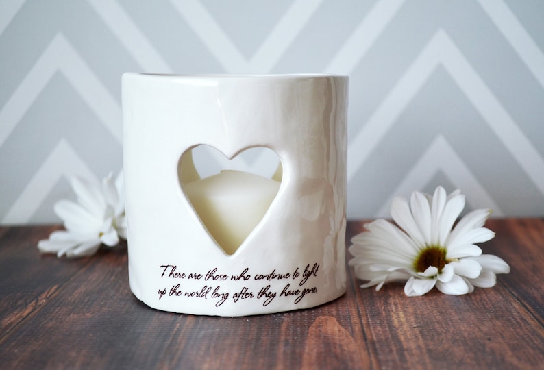 Product image of an off white ceramic votive with a white tea candle inside. The candle is visible through a heart shaped cutout on the front and back. The votive has two lines of centered light brown sepia text below the heart on the front.