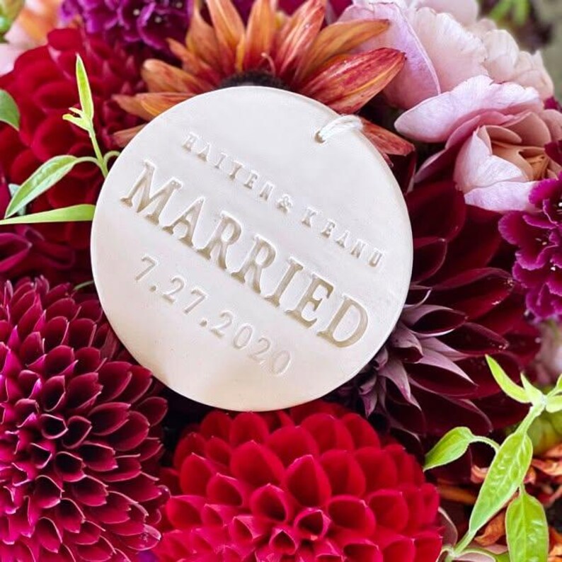 May include: A circular white ceramic ornament featuring the embossed text "HAITEN & KEANU", "MARRIED", and the date "7.27.2020". The ornament has a small hole at the top for hanging and is positioned against a background of deep red and pink flowers.