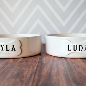 2 round off-white gloss-glazed large ceramic dog bowls are shown sitting on a wooden table. Each bowl has a dog bone shaped tile, stamped with a pet name, attached to the front. The stamped text is in all capitals, centered, and painted in black.