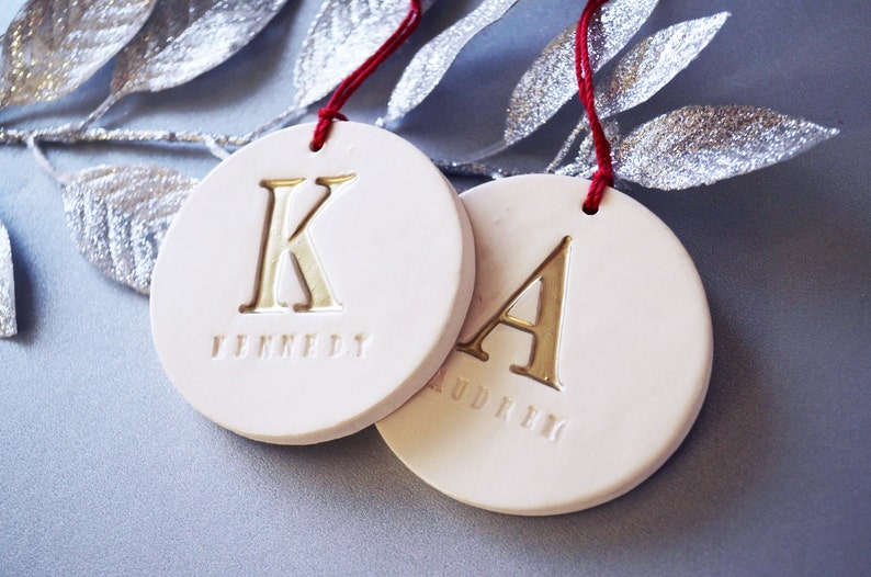 Close up of two off-white, round, ceramic ornaments. Each ornament is stamped with a large initial painted in metallic gold. A small name is stamped below the initial. Each ornament has a red cord to hang it from.