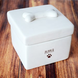 K&ouml;nnte beinhalten: Wei&szlig;e Keramikbox mit einem knochenf&ouml;rmigen Deckel und dem Namen "Pepper" auf der Vorderseite mit einem Pfotenabdruck.