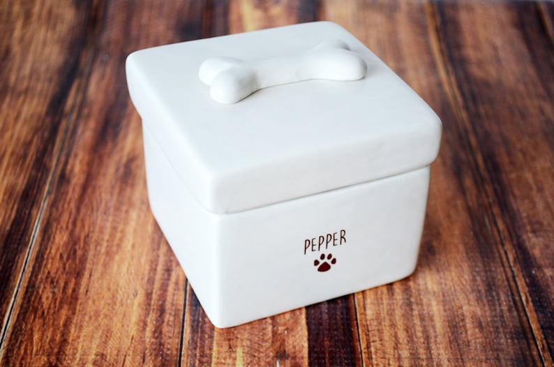 K&ouml;nnte beinhalten: Wei&szlig;e Keramikbox mit einem knochenf&ouml;rmigen Deckel und dem Namen "Pepper" auf der Vorderseite mit einem Pfotenabdruck.
