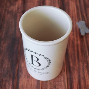 May include: White ceramic utensil holder with a black floral wreath design and the letter "B" in the center. The text "Jamie & Mark" and "Est. 2019" are printed below the wreath.