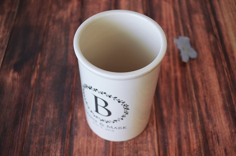 May include: White ceramic utensil holder with a black floral wreath design and the letter "B" in the center. The text "Jamie & Mark" and "Est. 2019" are printed below the wreath.