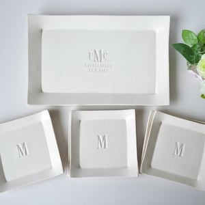 May include: A set of white ceramic serving dishes and plates. The set includes a large rectangular tray with embossed initials and a date, and three square plates with a single initial. A bouquet of flowers is in the background.