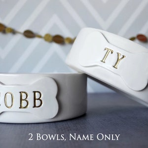 Image of the 2 bowls, name only option. Two bowls are shown, each with a bone shaped tile on the front. Each tile is stamped with a different pet name in all capital letters and painted in metallic gold.