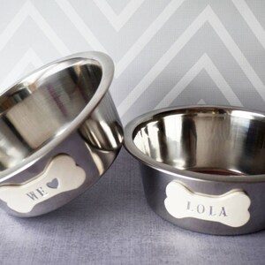 May include: Two stainless steel pet bowls. One bowl has the word "WE" on a bone-shaped tag, and the other has "LOLA". The bowls are set against a gray and white chevron pattern background.