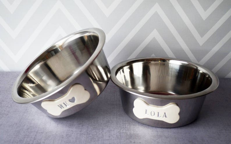 May include: Two stainless steel pet bowls. One bowl has the word "WE" on a bone-shaped tag, and the other has "LOLA". The bowls are set against a gray and white chevron pattern background.