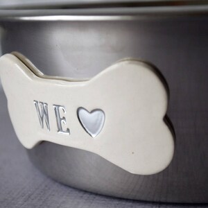 May include: A stainless steel pet bowl with a bone-shaped accent. The bone is off-white with the word "WE" and a heart symbol. The bowl has a shiny, metallic finish.