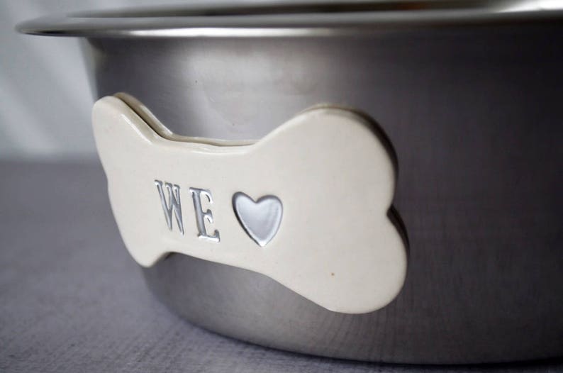 May include: A stainless steel pet bowl with a bone-shaped accent. The bone is off-white with the word "WE" and a heart symbol. The bowl has a shiny, metallic finish.