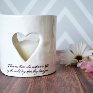 Product image of an off white ceramic votive with a white tea candle inside. The candle is visible through a heart shaped cutout on the front and back. The votive has two lines of centered light brown sepia text below the heart on the front.