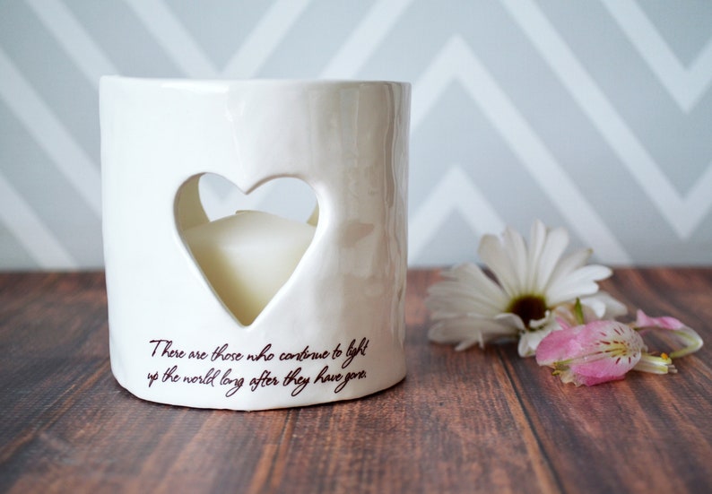 Product image of an off white ceramic votive with a white tea candle inside. The candle is visible through a heart shaped cutout on the front and back. The votive has two lines of centered light brown sepia text below the heart on the front.