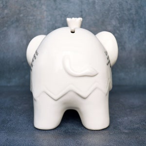 Peut inclure: Une tirelire blanche en forme d'&eacute;l&eacute;phant en c&eacute;ramique. La tirelire a une fente sur le dessus et une petite couronne. Les oreilles de l'&eacute;l&eacute;phant sont visibles, et il y a du texte sur le c&ocirc;t&eacute;. Le bas de la tirelire a un motif en zigzag.