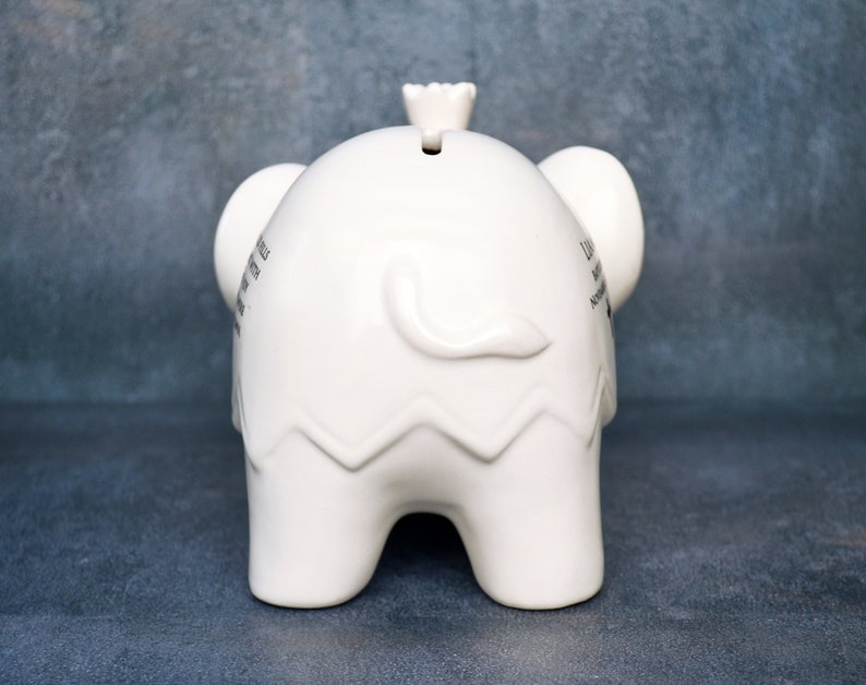 Peut inclure: Une tirelire blanche en forme d'&eacute;l&eacute;phant en c&eacute;ramique. La tirelire a une fente sur le dessus et une petite couronne. Les oreilles de l'&eacute;l&eacute;phant sont visibles, et il y a du texte sur le c&ocirc;t&eacute;. Le bas de la tirelire a un motif en zigzag.