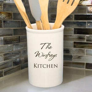 May include: A cream-coloured kitchen utensil holder with the text "The Winfreys KITCHEN" in black script. The holder contains wooden spoons and a grey spatula. The background features a mosaic tile backsplash.