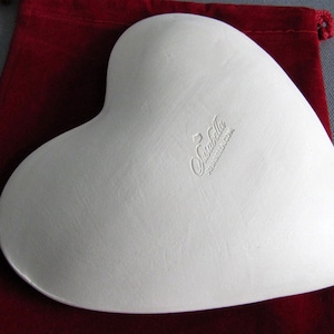 May include: White heart-shaped ceramic dish with a raised rim. The dish is made by "Sunbella" and has a "Made in China" label on the bottom.