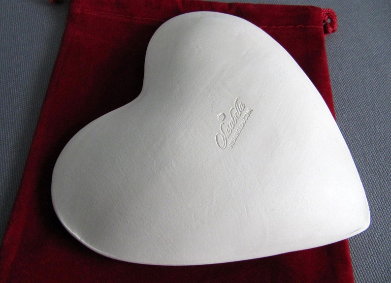 May include: White heart-shaped ceramic dish with a raised rim. The dish is made by "Sunbella" and has a "Made in China" label on the bottom.