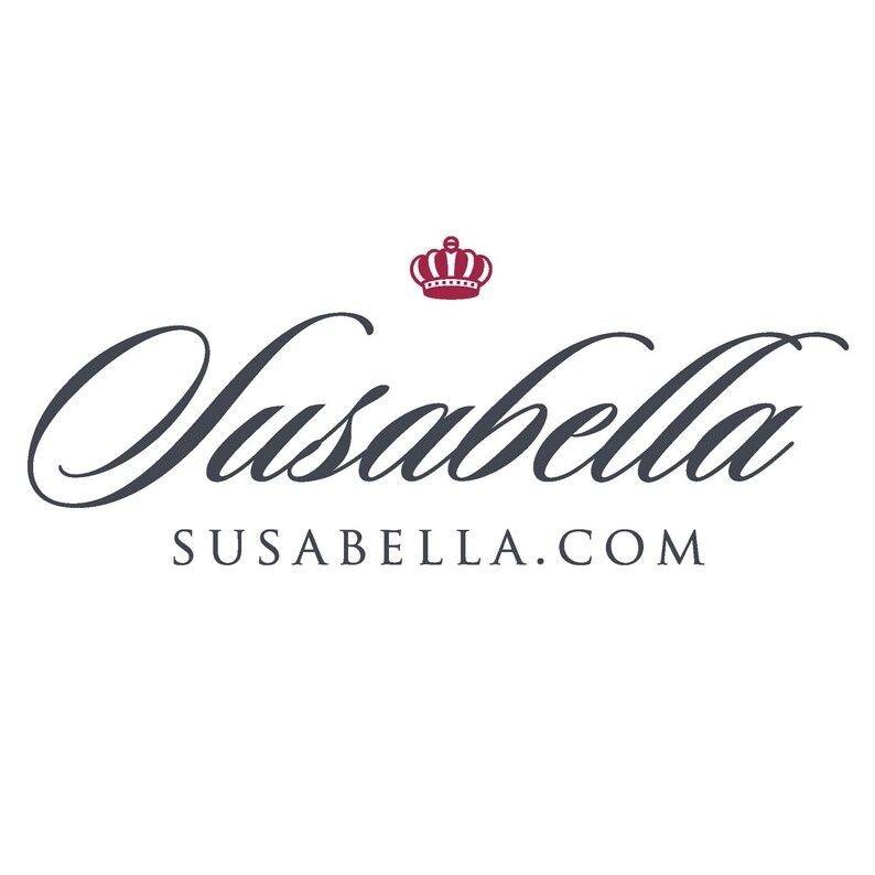 May include: A logo for a company called Susabella. The logo features a stylized cursive font with the company name "Susabella" in dark gray. The name is centered above the company website address "susabella.com" in a smaller font. A small red crown is positioned above the company name.