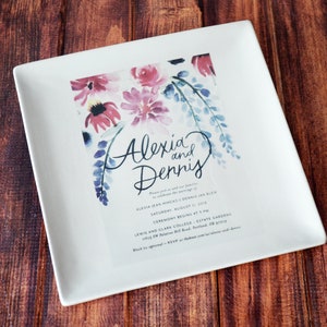 A large square serving plate sits on a wooden table. The plate has a full color custom image centered on the front. The image is a custom wedding invitation with flowers at the top and text. The plate is white with a gloss glaze finish.