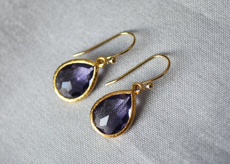 K&ouml;nnte beinhalten: Ein Paar goldene Tropfenohrringe mit violetten Edelstein-Akzenten.
