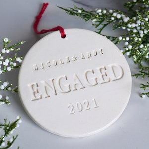 Product image of an off white gloss glazed ceramic round shaped  ornament. The front is stamped with custom text in all capital letters. The text is centered and reads Nicole & Andy, Engaged, 2021. The ornament has a red cord to hang from.