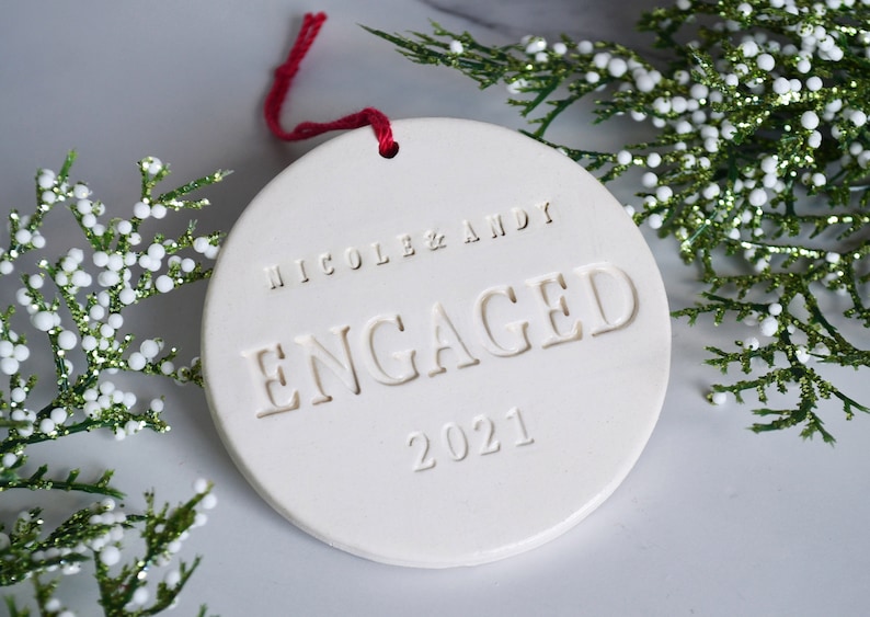 Product image of an off white gloss glazed ceramic round shaped  ornament. The front is stamped with custom text in all capital letters. The text is centered and reads Nicole & Andy, Engaged, 2021. The ornament has a red cord to hang from.