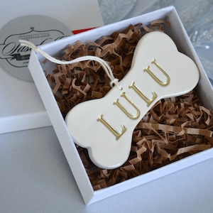 An off-white gloss-glazed ceramic, dog bone-shaped ornament with the pet name "LULU" stamped into the surface in all capital letters. The name is painted in metallic gold. The ornament has a white cord and is shown in a white gift box.