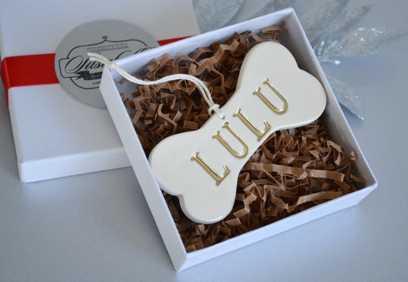 An off-white gloss-glazed ceramic, dog bone-shaped ornament with the pet name "LULU" stamped into the surface in all capital letters. The name is painted in metallic gold. The ornament has a white cord and is shown in a white gift box.