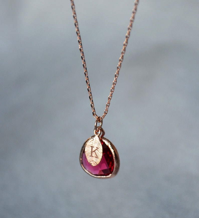 A pinkish red ruby colored transparent glass stone charm is framed in rose gold metal and hangs from a rose gold link chain. There is a small leaf shaped charm with the letter K stamped on the front, hanging in front of the ruby charm.