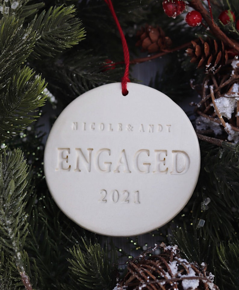 Product image of an off white gloss glazed ceramic round shaped ornament. The front is stamped with custom text in all capital letters. The text is centered and reads Nicole & Andy, Engaged, 2021. The ornament has a red cord to hang from.