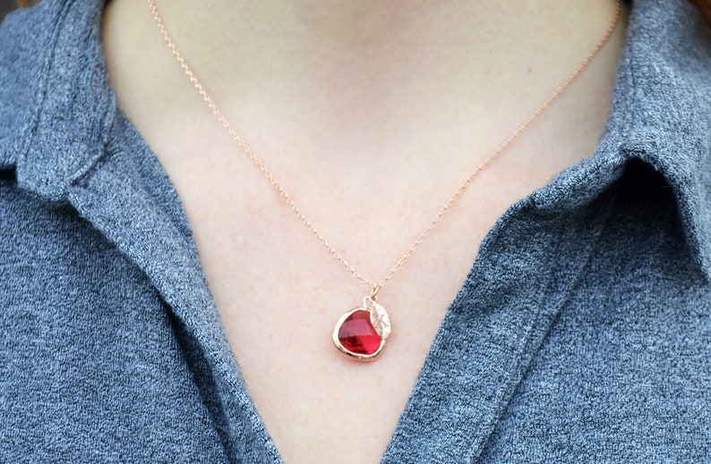 The necklace is shown on a model wearing a grey button down shirt. A rose gold metal chain with ruby and leaf charm hangs around the model's neck. The ruby is framed in rose gold and the leaf on top is stamped with a custom initial.