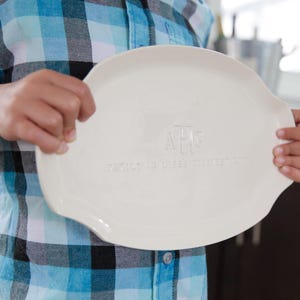 May include: A white ceramic oval platter with a monogram and text. The platter is held by a person wearing a blue and black plaid shirt. The platter has a slightly raised edge and is smooth.