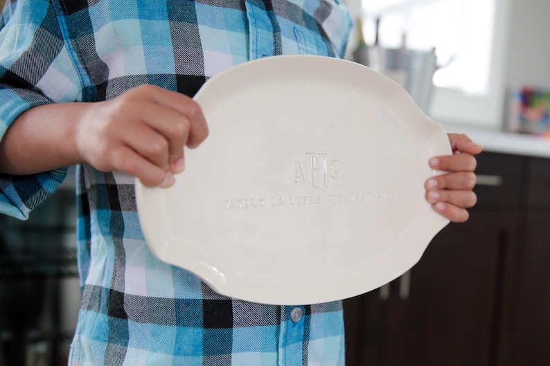 May include: A white ceramic oval platter with a monogram and text. The platter is held by a person wearing a blue and black plaid shirt. The platter has a slightly raised edge and is smooth.