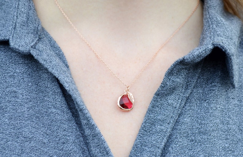 The necklace is shown on a model wearing a grey button down shirt. A rose gold metal chain with ruby and leaf charm hangs around the model's neck. The ruby is framed in rose gold and the leaf on top is stamped with a custom initial.