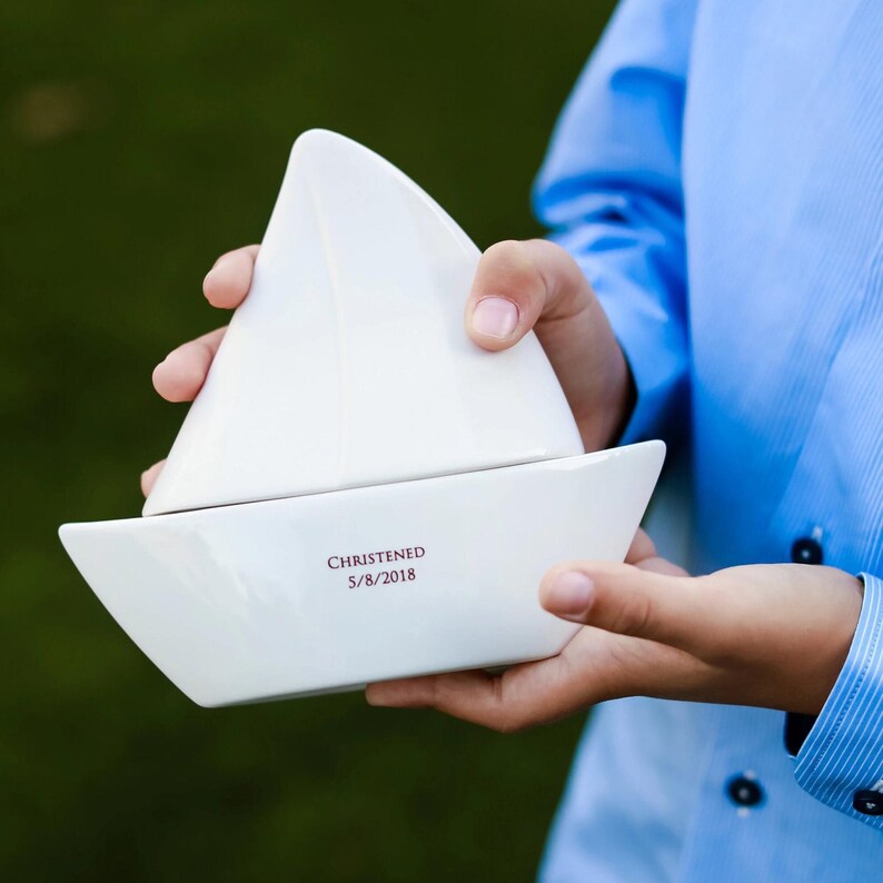 Op de afbeelding: Een witte keramische keepsake box in de vorm van een zeilboot. De doos wordt vastgehouden door een persoon die een blauw overhemd draagt. De tekst "CHRISTENED 5/8/2018" is in het rood op de basis van de boot gedrukt. De zeilboot heeft een driehoekig zeil en een rechthoekige basis.