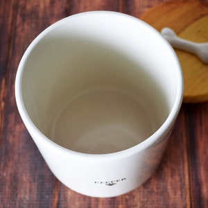 Puede incluir: Tarro de cer&aacute;mica blanco con tapa de madera con un pomo en forma de hueso. El tarro tiene la palabra "Pup-per" impresa en la parte inferior.