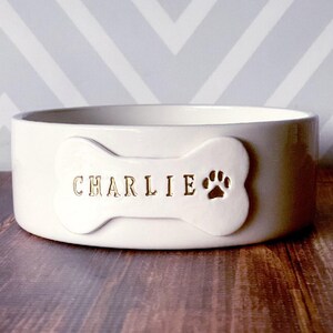 May include: A round, off-white ceramic pet bowl featuring a raised bone-shaped plaque on the front. The plaque is embossed with the name CHARLIE in gold lettering, followed by a gold-coloured paw print icon centred on the right side of the bone.