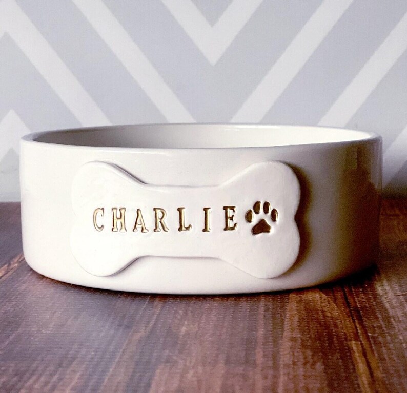 May include: A round, off-white ceramic pet bowl featuring a raised bone-shaped plaque on the front. The plaque is embossed with the name CHARLIE in gold lettering, followed by a gold-coloured paw print icon centred on the right side of the bone.