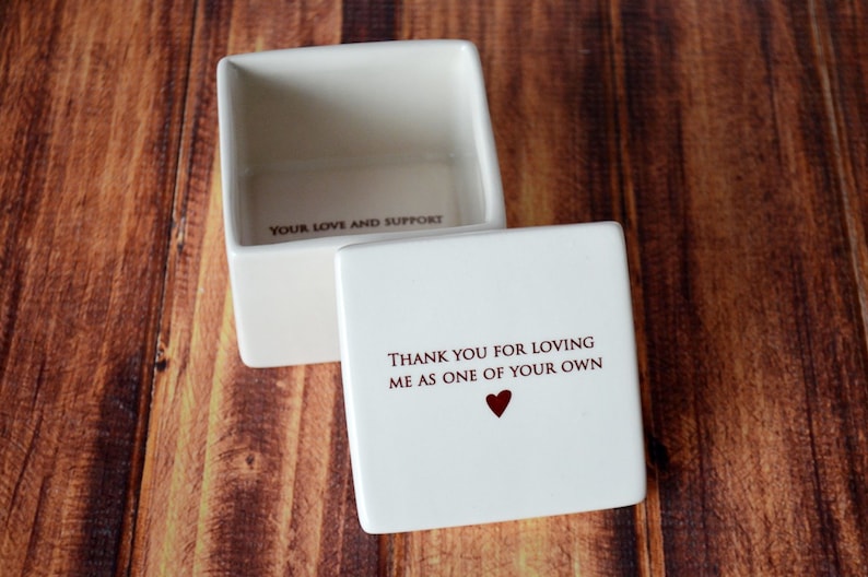 May include: A white square ceramic keepsake box with the lid slightly ajar. The lid has the text "THANK YOU FOR LOVING ME AS ONE OF YOUR OWN" with a red heart. The box interior reads "YOUR LOVE AND SUPPORT".
