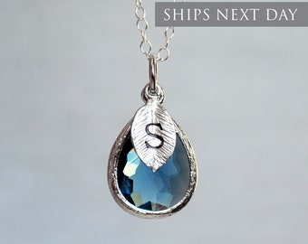 May include: A silver necklace with a teardrop-shaped blue crystal pendant. The pendant has a silver leaf charm with the letter "S" engraved on it.