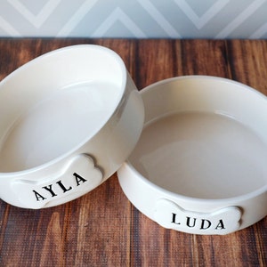 2 round off-white gloss-glazed large ceramic dog bowls are shown sitting on a wooden table. A top view shows the circular shape, wide with straight sides, creating a shallow bowl for food or water. Each bowl has a bone-shaped tile on the front.