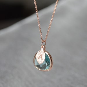 May include: Rose gold necklace with a teardrop-shaped, light blue gemstone pendant. A small leaf charm with the letter "A" is attached to the pendant. The chain is delicate and the background is blurred.