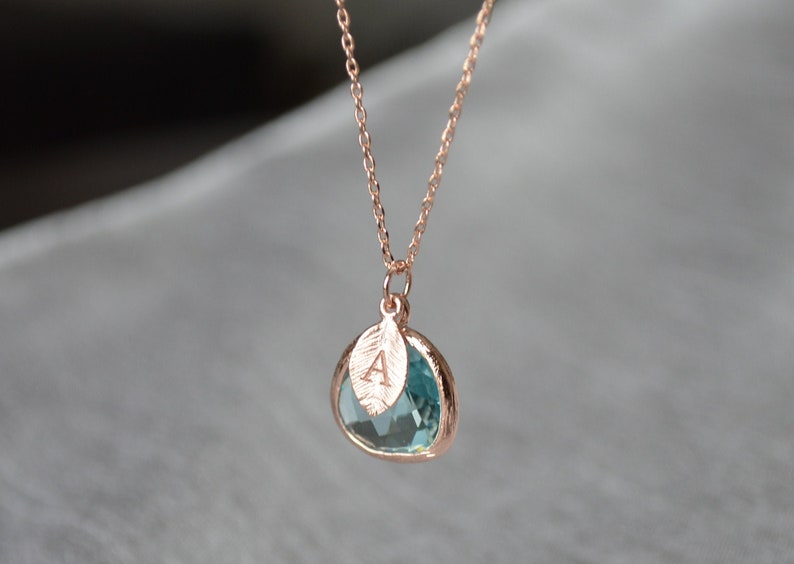 May include: Rose gold necklace with a teardrop-shaped, light blue gemstone pendant. A small leaf charm with the letter "A" is attached to the pendant. The chain is delicate and the background is blurred.