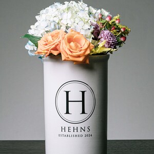 Puede incluir: Un jarr&oacute;n de cer&aacute;mica blanco con un arreglo floral. El jarr&oacute;n tiene un logotipo circular negro con la letra "H" y el texto "HEHNS ESTABLISHED 2024". Las flores incluyen rosas naranjas, hortensias azules y blancas y otras flores coloridas.