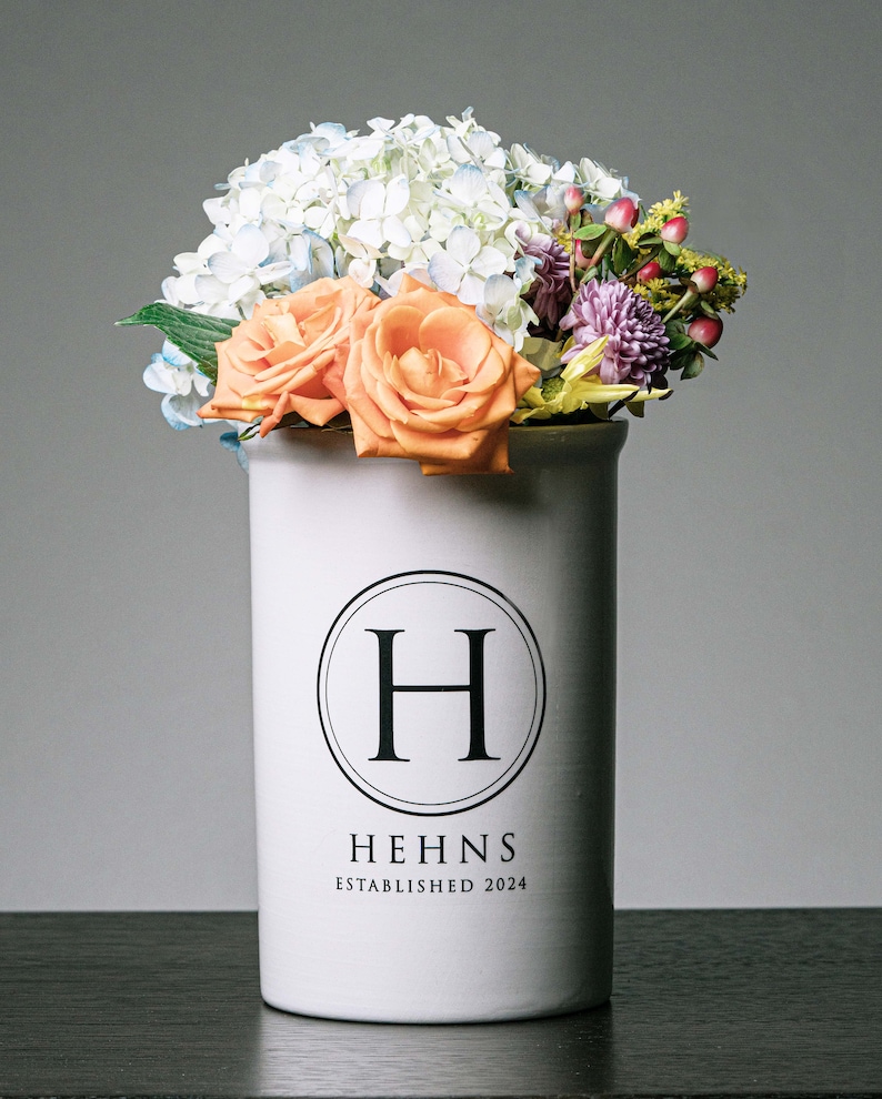 Puede incluir: Un jarr&oacute;n de cer&aacute;mica blanco con un arreglo floral. El jarr&oacute;n tiene un logotipo circular negro con la letra "H" y el texto "HEHNS ESTABLISHED 2024". Las flores incluyen rosas naranjas, hortensias azules y blancas y otras flores coloridas.