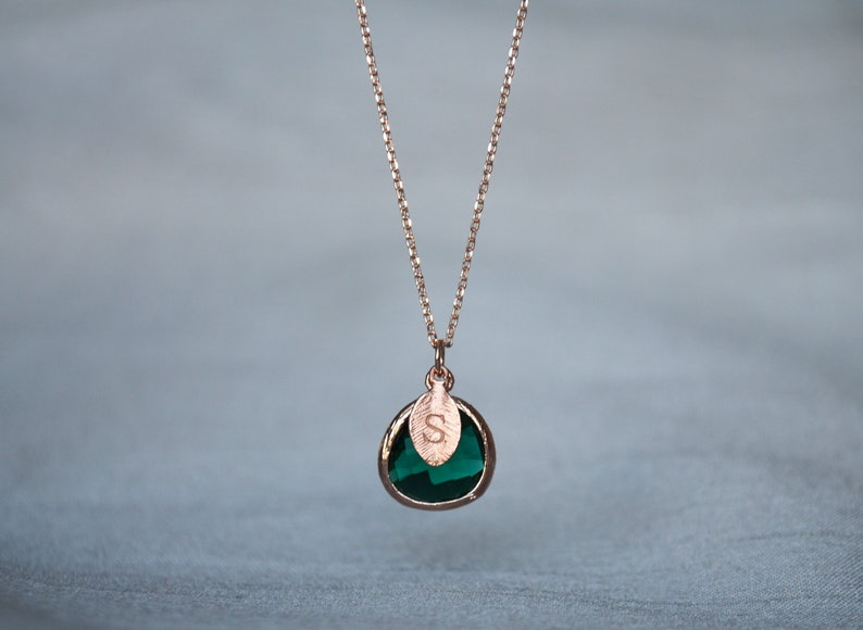 A green emerald colored transparent glass stone charm is framed in rose gold metal and hangs from a rose gold link chain. There is a small leaf shaped charm with the letter S stamped on the front, hanging in front of the emerald charm.