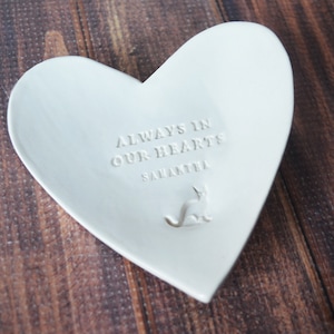 May include: White ceramic heart-shaped dish with the text "Always in our hearts" and the name "Samantha" engraved on the front. A small cat silhouette is also engraved on the dish.