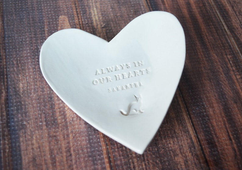 May include: White ceramic heart-shaped dish with the text "Always in our hearts" and the name "Samantha" engraved on the front. A small cat silhouette is also engraved on the dish.
