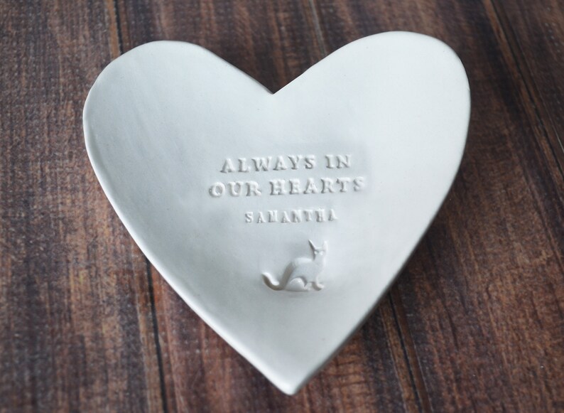 May include: White ceramic heart-shaped dish with the inscription "Always in our hearts Samantha" and a small cat silhouette.