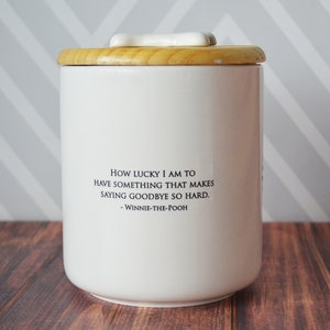 Puede incluir: Tarro de cer&aacute;mica blanco con tapa de madera y pomo en forma de hueso. El tarro tiene una cita de Winnie de Pooh que dice: "Qu&eacute; suerte tengo de tener algo que hace que decir adi&oacute;s sea tan dif&iacute;cil. - Winnie de Pooh"